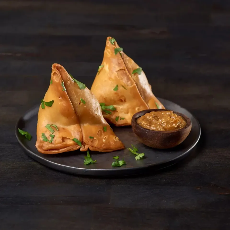 Two golden samosas garnished with cilantro, accompanied by a bowl of dipping sauce on a dark plate.