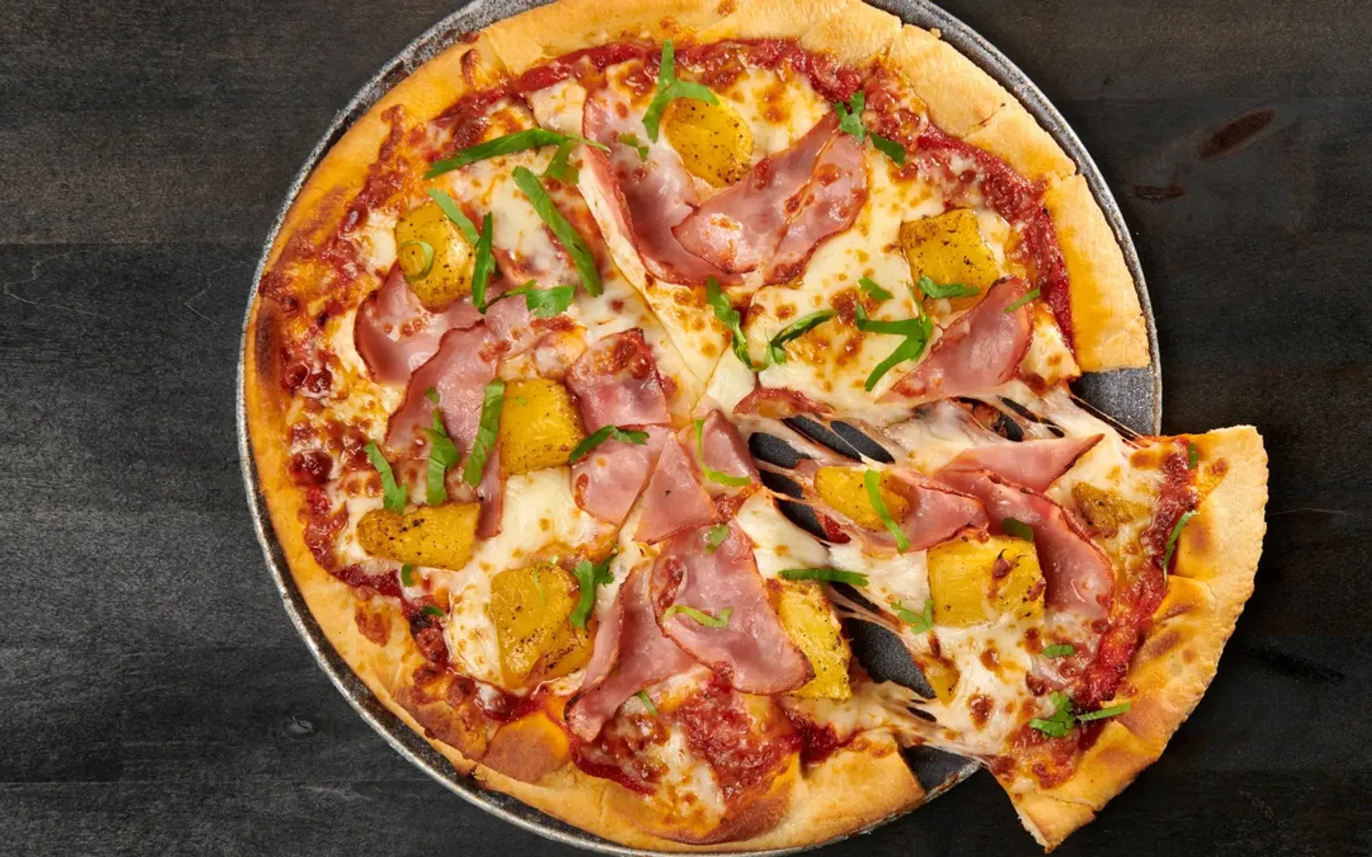 Cheesy pizza topped with ham, pineapple, and green herbs, with a slice being pulled away.