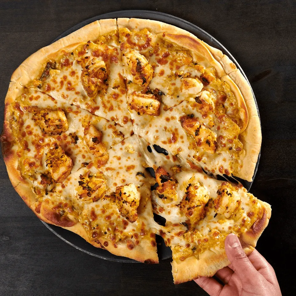 A hand reaches for a slice of cheesy pizza topped with golden-brown chicken pieces.