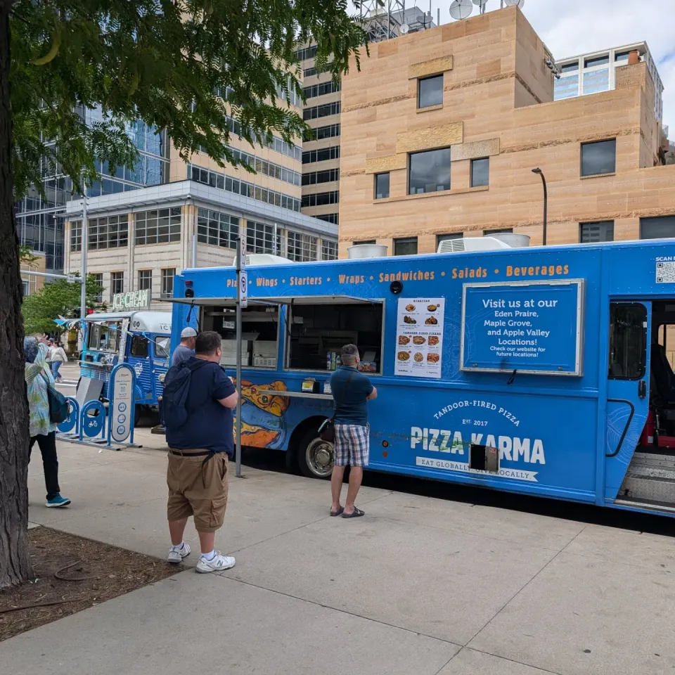 A blue pizza food truck with a menu, while customers wait in line to order.