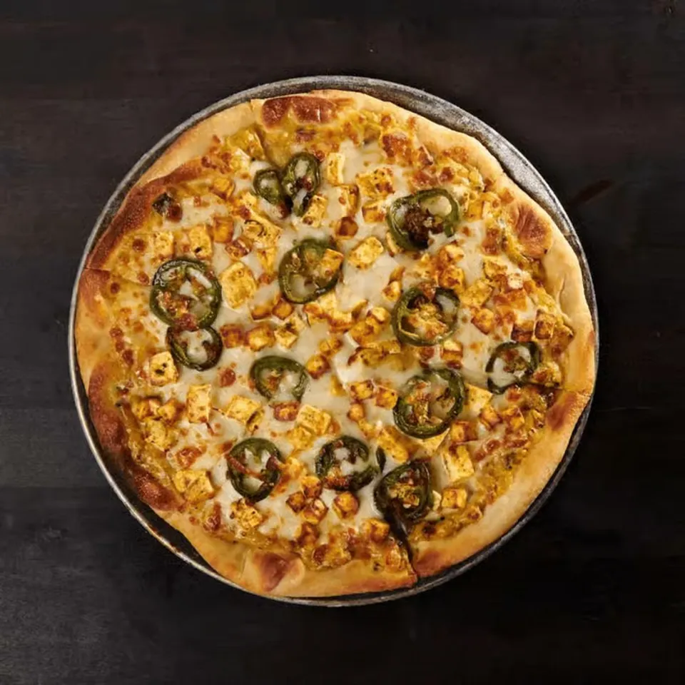 Crispy pizza topped with cheese, paneer, jalapeños, and a golden-brown crust on a wooden surface.
