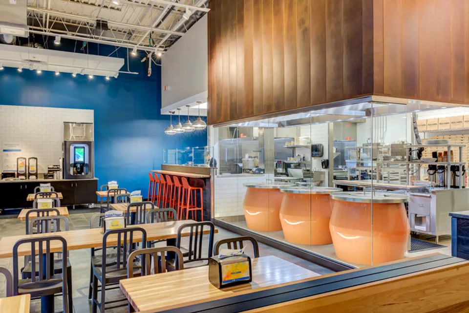 Bright, modern restaurant interior with wooden tables, red bar stools, and an open kitchen view.