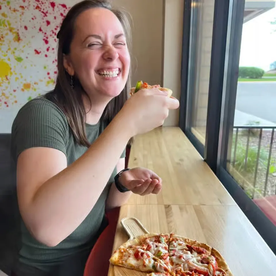 Smiling woman enjoying a slice of pizza at a wooden table by a large window.
