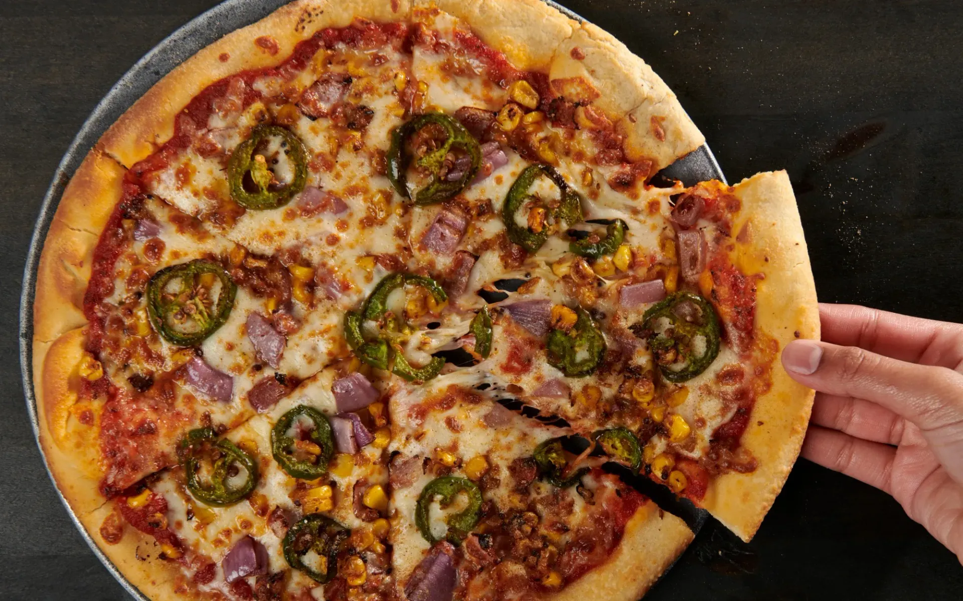 A hand reaches for a slice of pizza topped with jalapeños, corn, and ham on a dark surface.