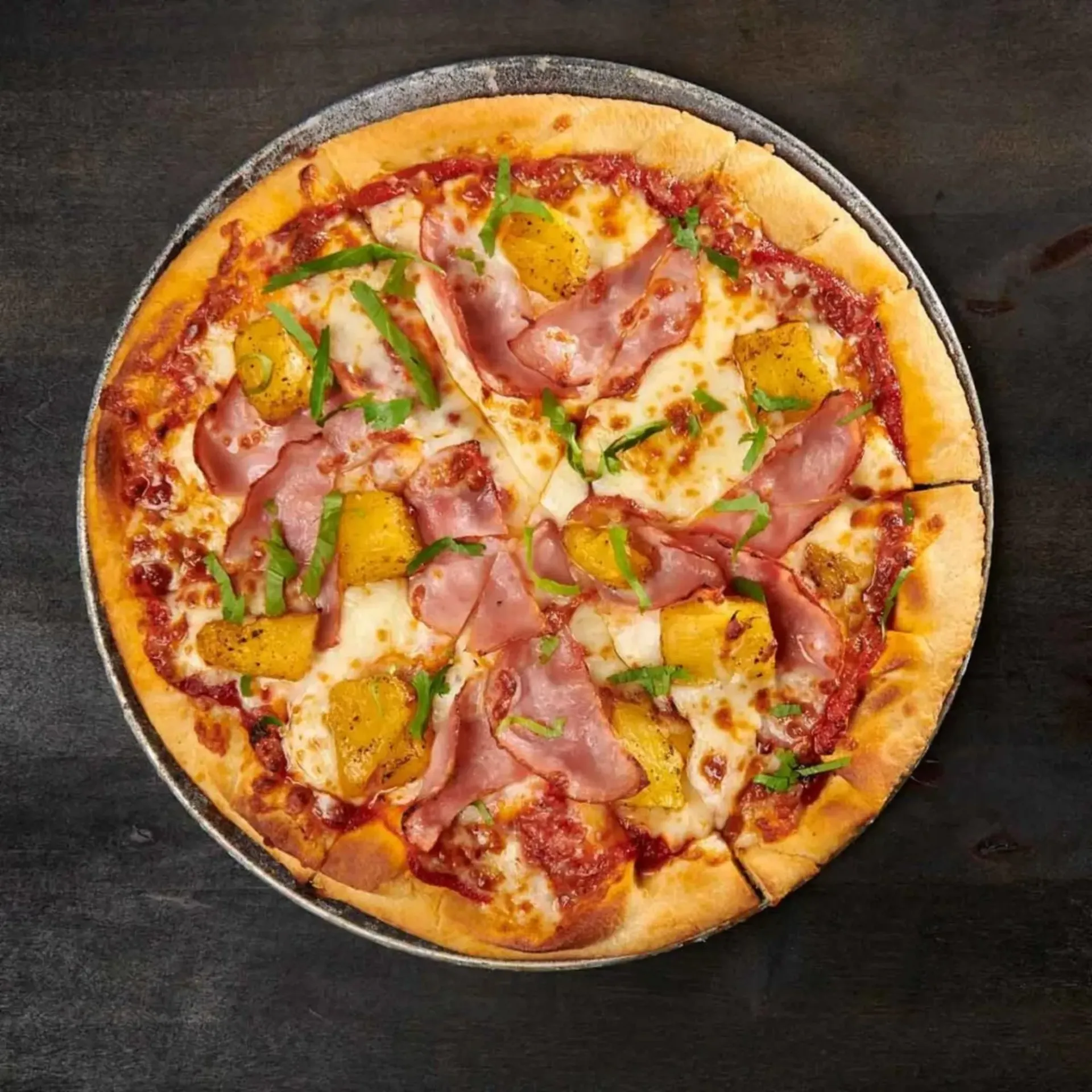 A pizza topped with ham, pineapple, cheese, and green herbs, served on a dark wooden surface.