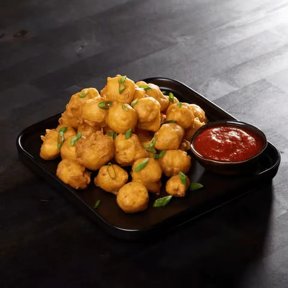 Golden, crispy fritters piled high on a black plate, garnished with green onions, accompanied by a bowl of red sauce.