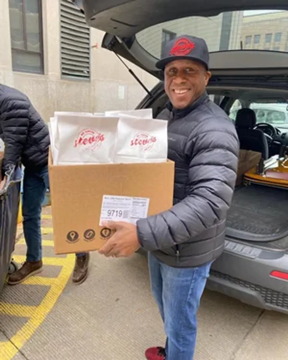 Smiling man in a black jacket and hat carries a box of food packages towards a vehicle.