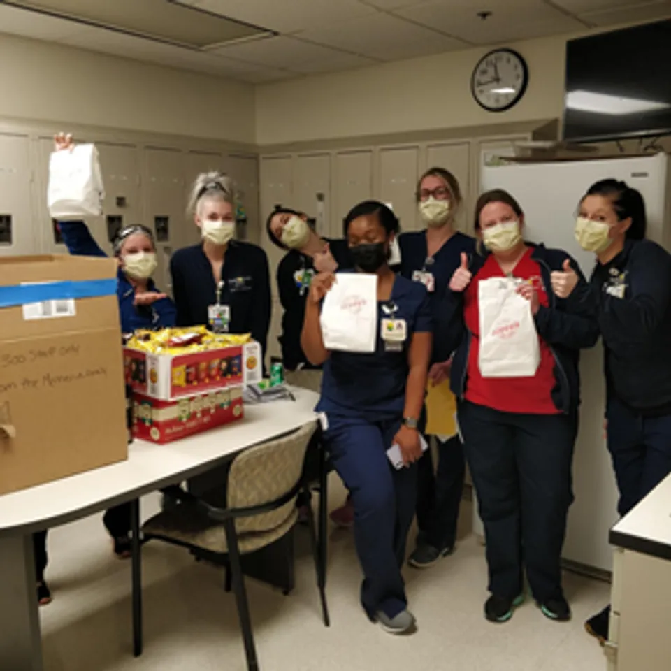 Healthcare workers in scrubs and masks celebrate with bags of food and snacks in a hospital break room.