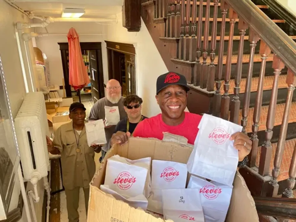 Four people stand on a staircase, one holding a box of takeout bags, smiling and engaging with the camera.