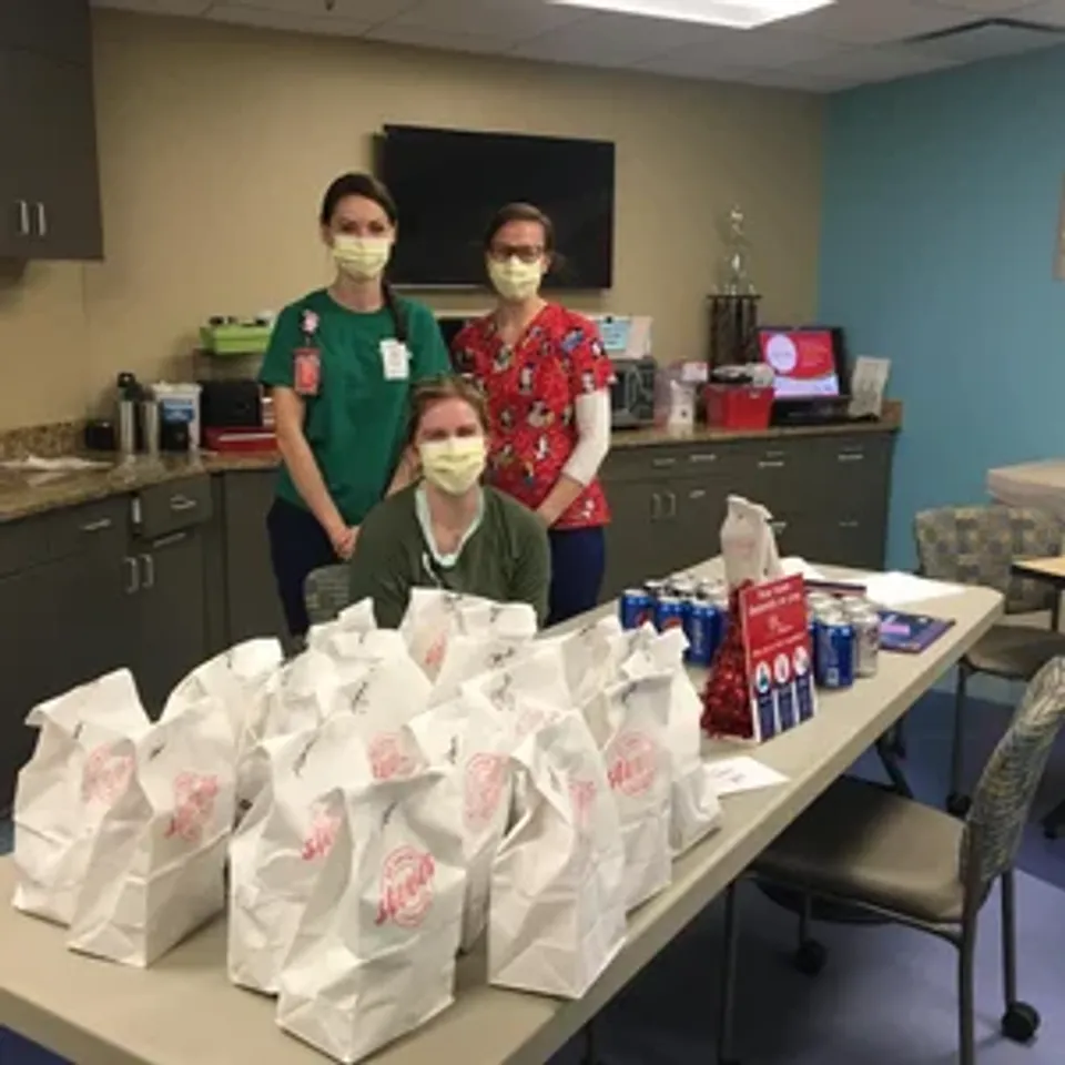 Three healthcare workers in masks stand behind a table filled with takeout bags and beverages in a break room.