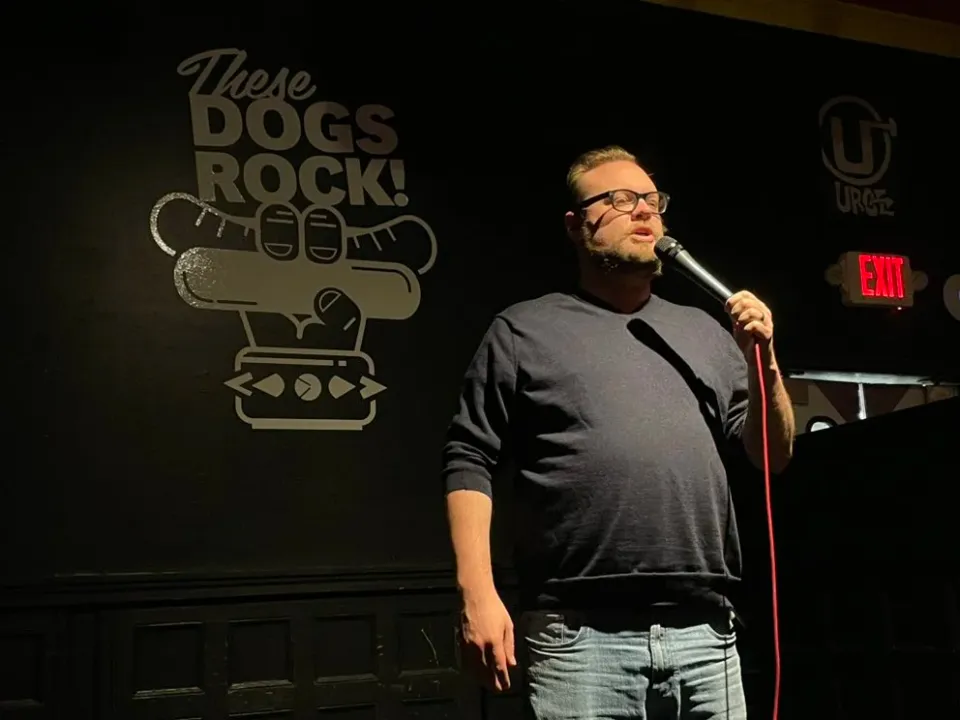 A man with a beard and glasses stands on stage delivering stand up comedy at open mic night at steve's hot doogs
