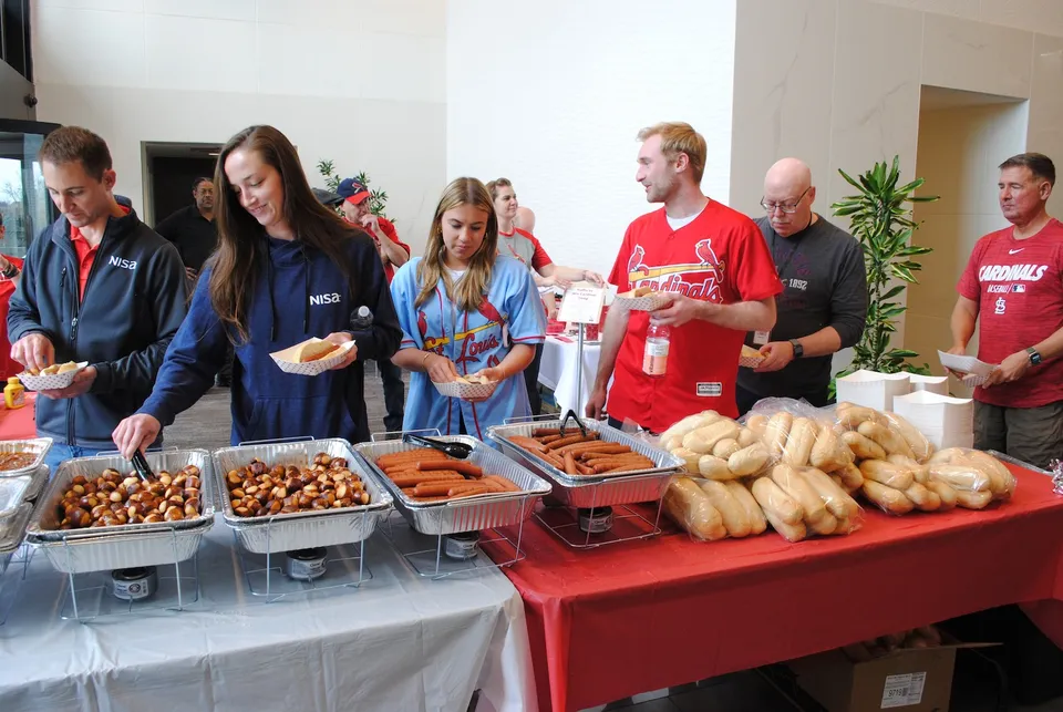 Group of people serving and enjoying food at a buffet, featuring sausages and bread rolls.
