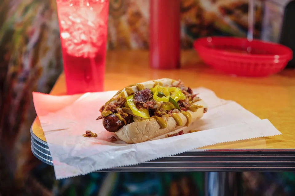 The St. Louis Style Hot Dog sits on a table with a drink behind it.