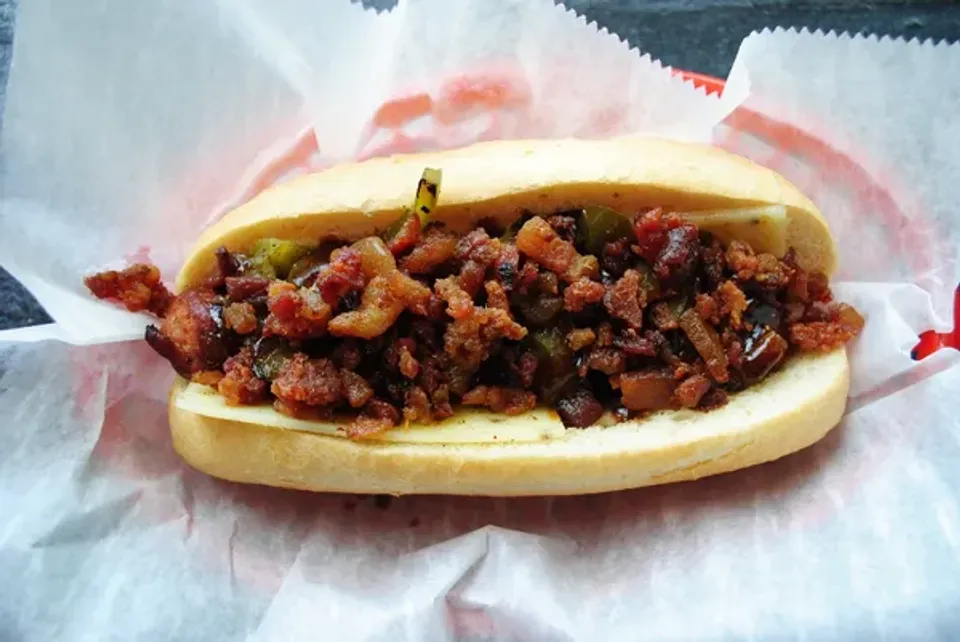 A hot dog in a bun topped with bacon green peppers, onions, pepper jack cheese