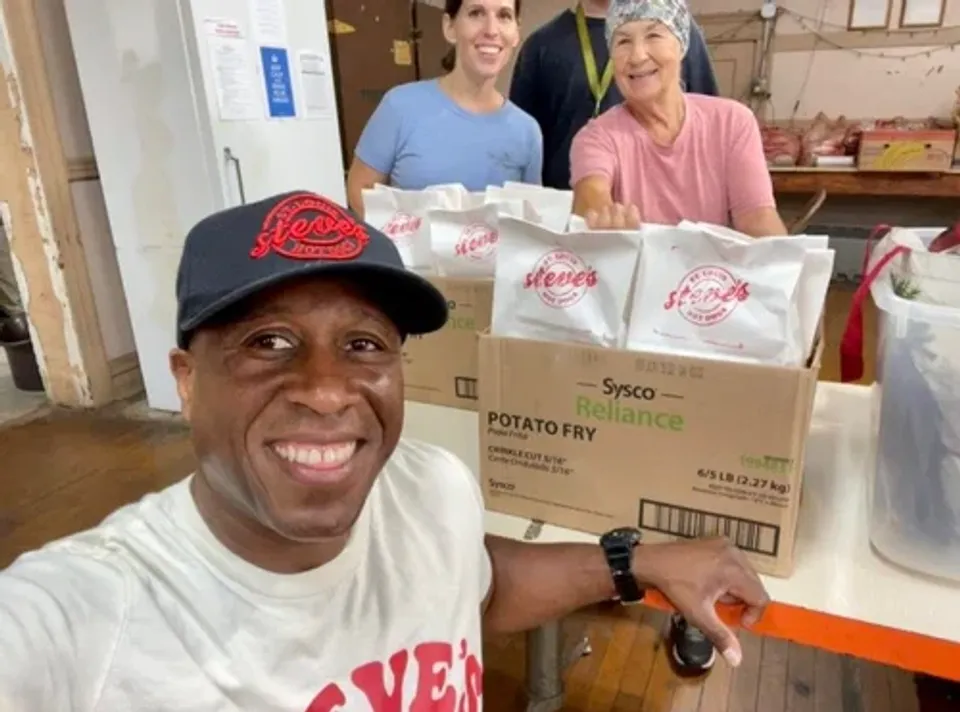 Three volunteers smile in a kitchen setting, with boxes of potato fry and supplies in the foreground.