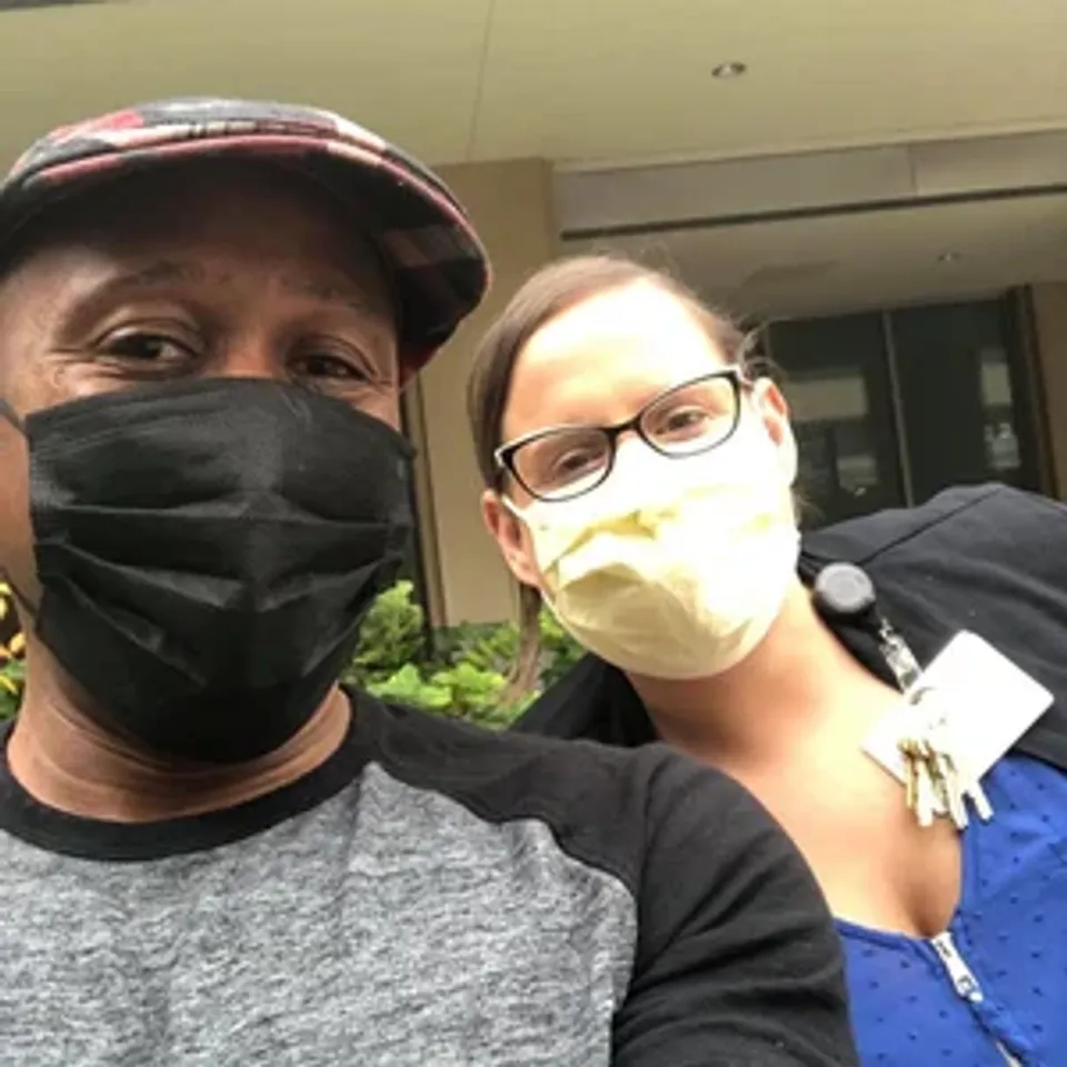 Two people wearing masks pose for a selfie outside a building, smiling at the camera.