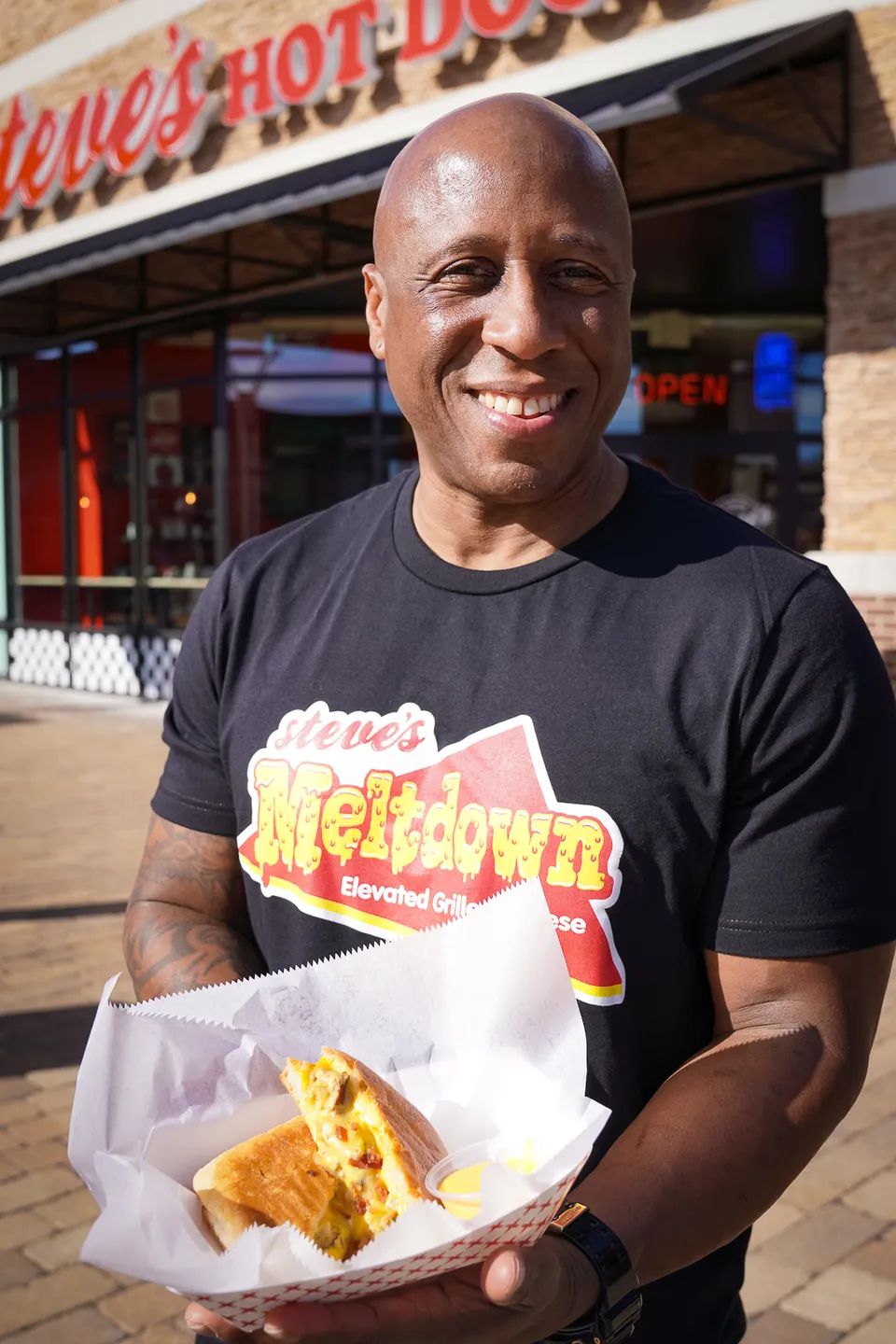 Steve Ewing stands outside Steve's Hot Dogs in a Steve's Meltdown shirt holding a grilled cheese sandwich