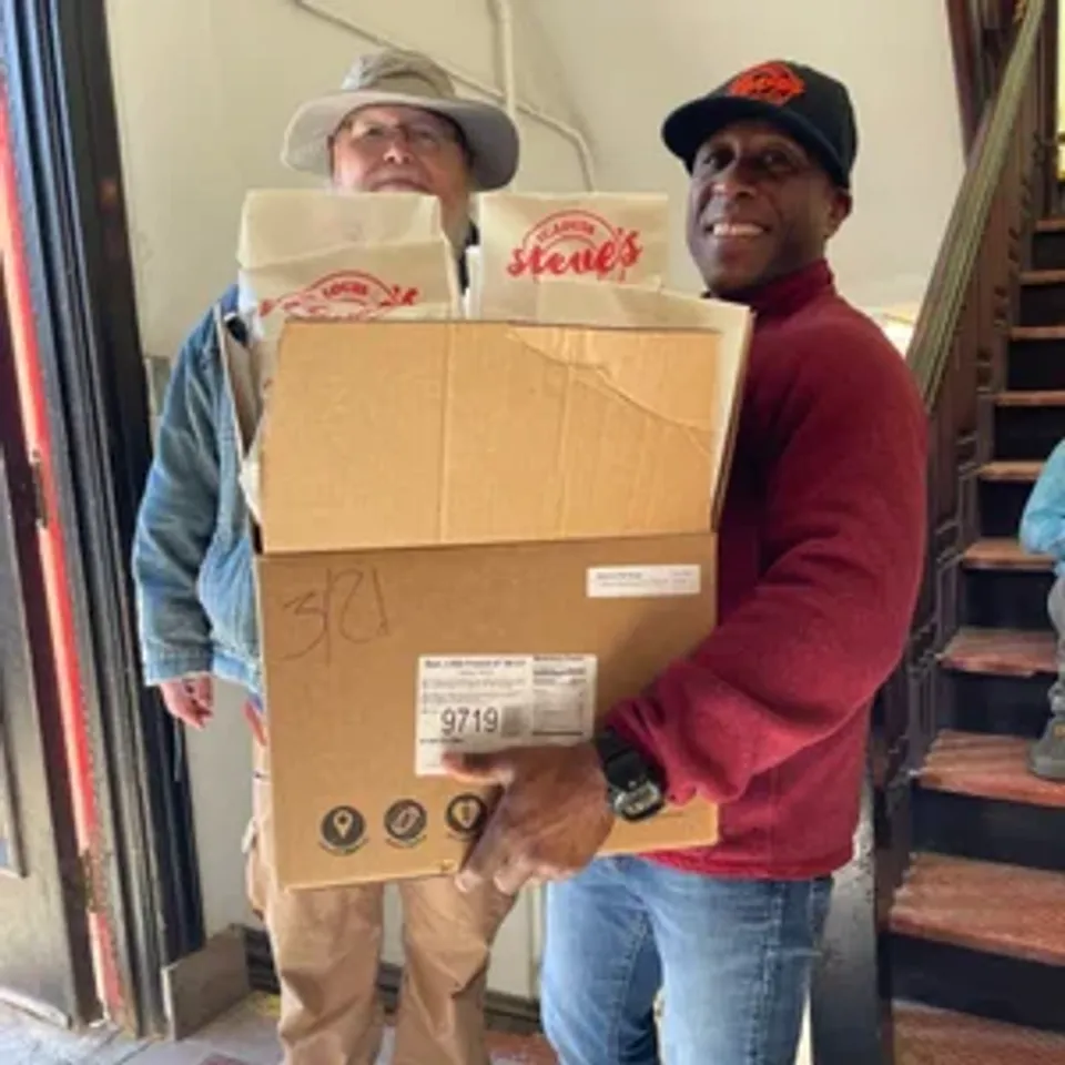 Two men stand indoors, one smiling while holding a large box filled with bags labeled "Stevie's."
