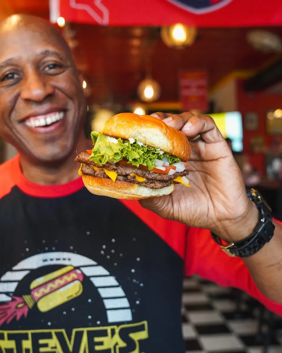 Steve Ewing Holds a Hey Now You're an All Star Burger with two all beef patties, topped with cheese, tomato, onion, pickle and ketchup