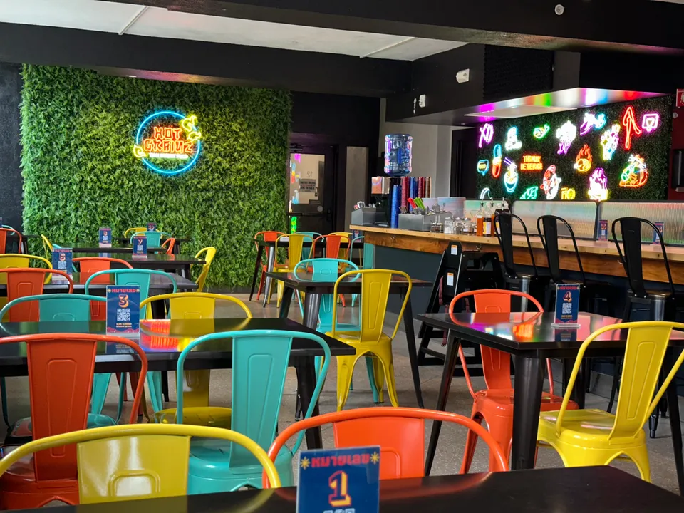 Colorful chairs surround tables in a vibrant cafe, featuring neon signage and greenery on the walls.